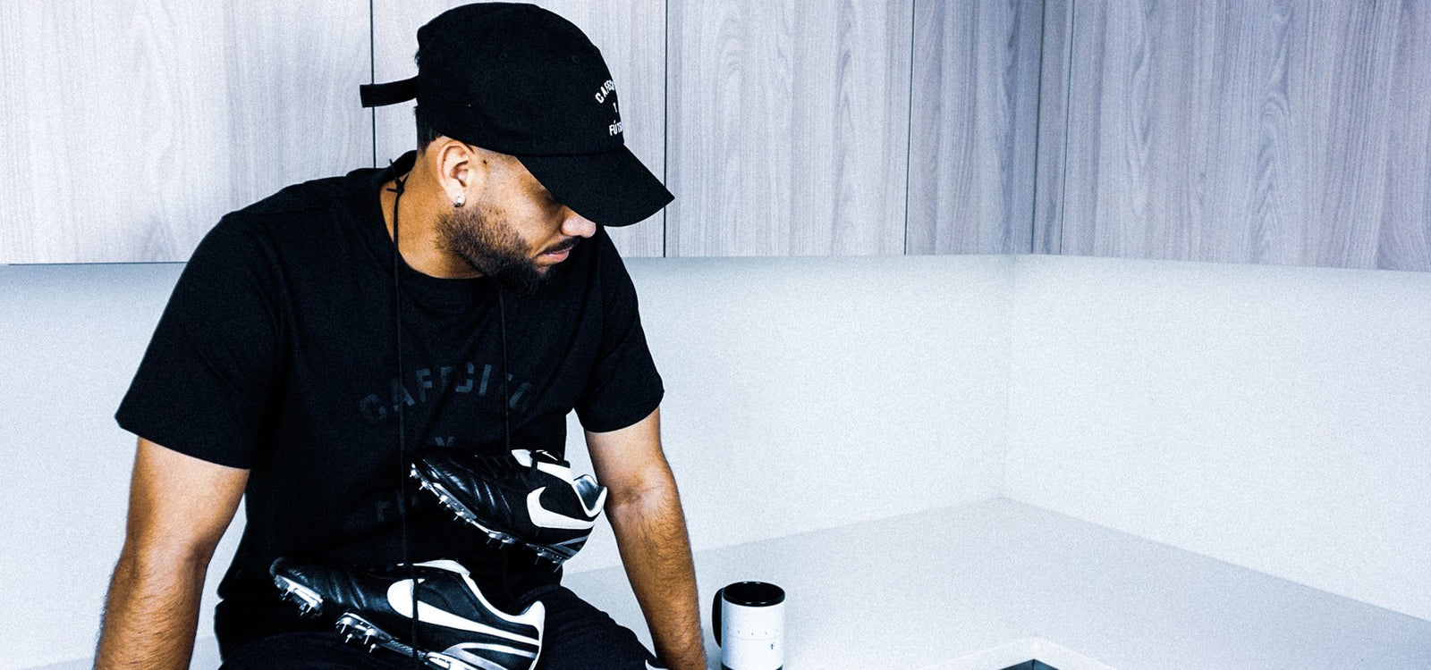 jesus gonzalez drinking coffee with nike tiempo legend cleats and cafecito y futbol t shirt in black and black coachs hat  