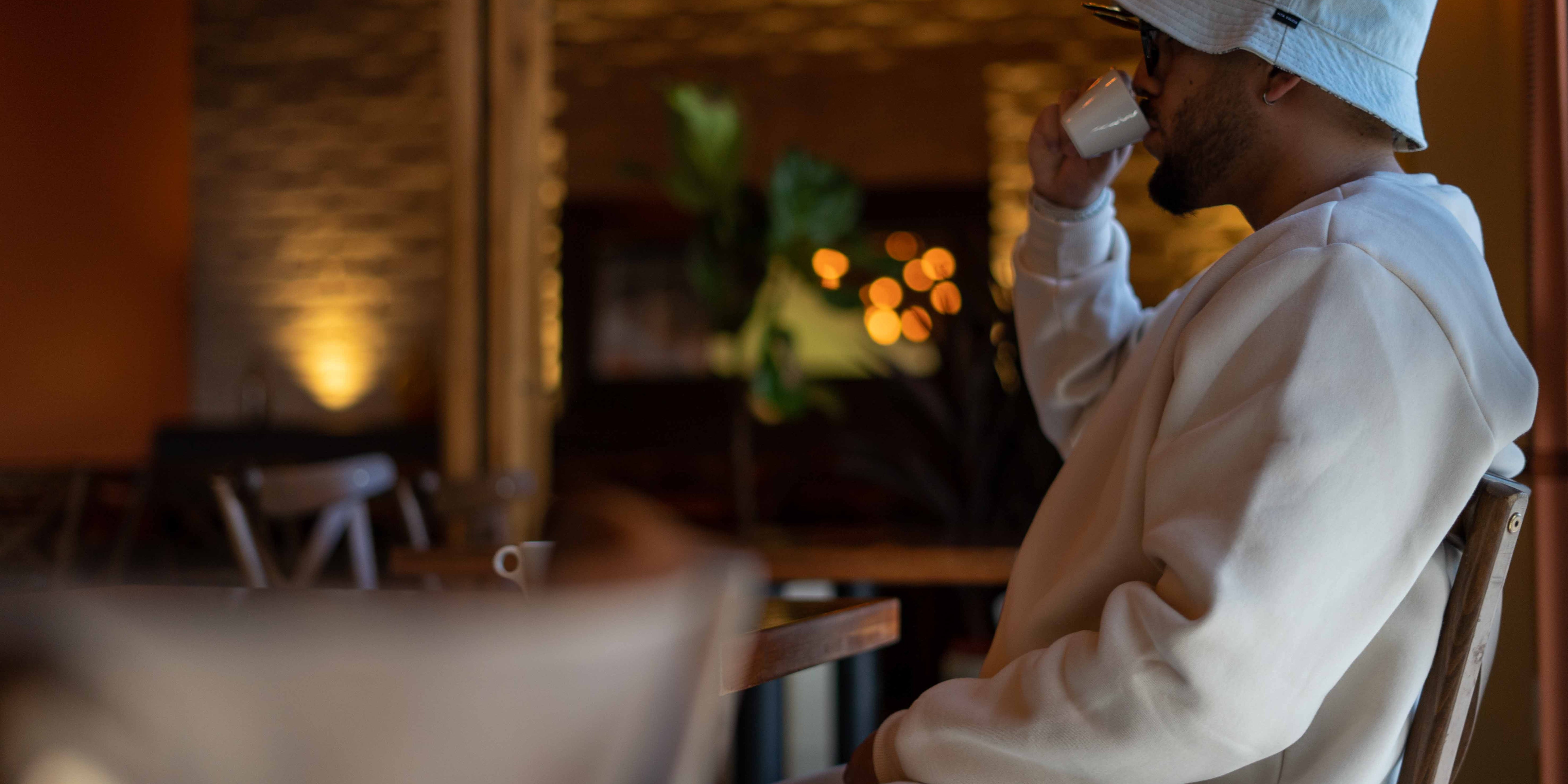 A man in a white bucket hat and matching white sweatshirt sits in a cafe setting, holding a white mug to his lips.