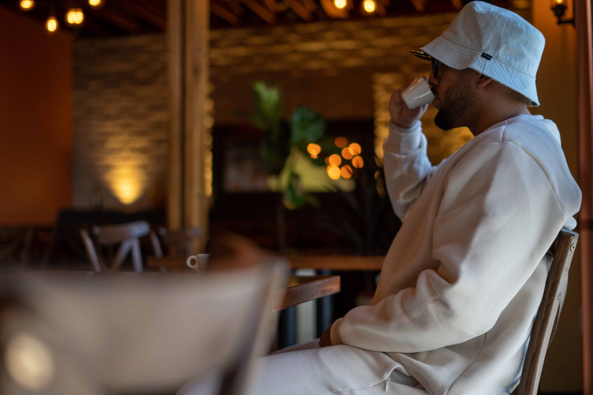 A man in a white bucket hat and matching white sweatshirt sits in a cafe setting, holding a white mug to his lips.