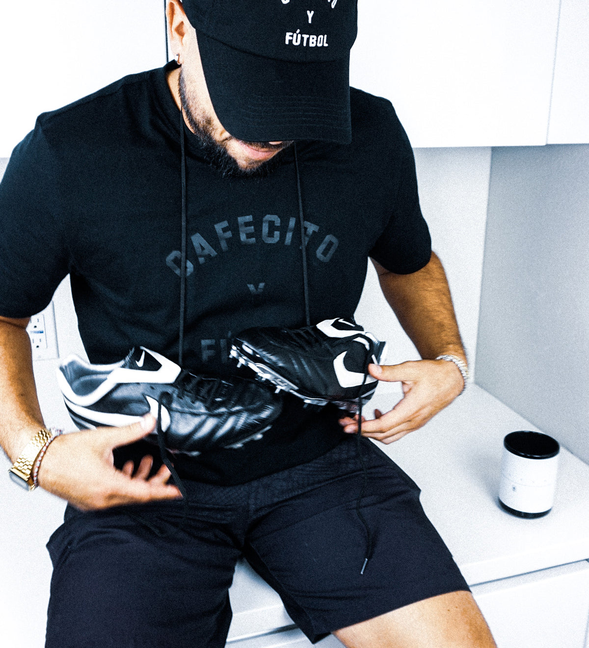 jesus gonzalez wearing cafecito y futbol coach's hat with cleats around his neck and cafecito y futbol t shirt in black on black
