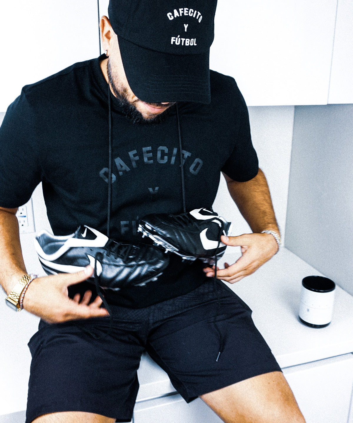 jesus gonzalez wearing cafecito y futbol coach's hat with cleats around his neck and cafecito y futbol t shirt in black on black