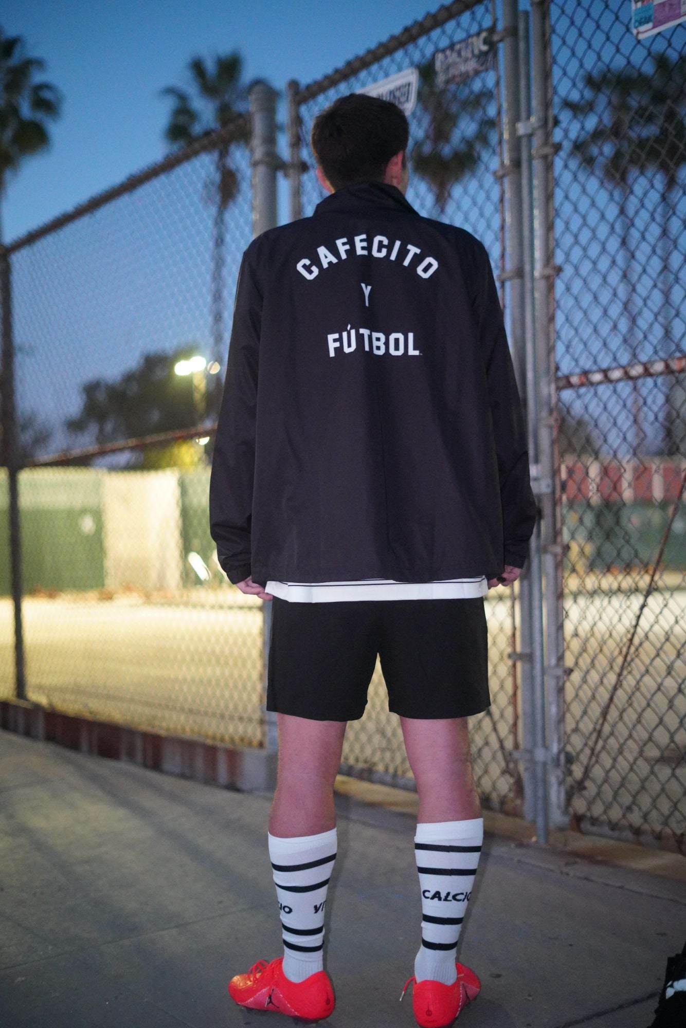 max rodriguez standing on pool side at pan pacific park by the grove at dusk facing away from the camera, wearing a black track jacket with 'CAFECITO Y FÚTbol' printed in white on the back, black shorts, white knee-high socks with black stripes, and bright red soccer cleats.